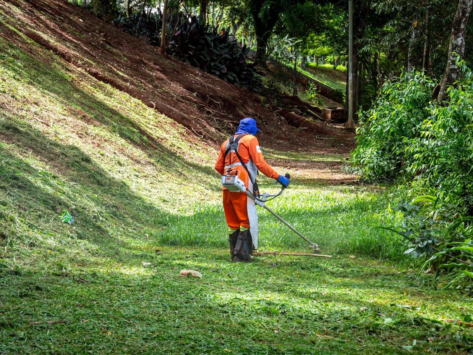 Un jardinier en tenue orange passe la débroussailleuse au pied d’un talus fraîchement dégagé